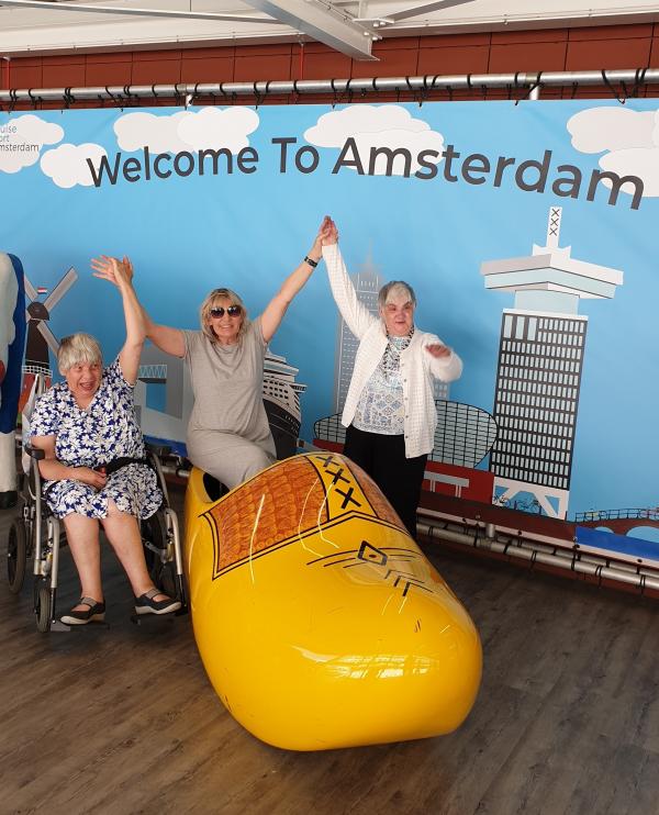 Three ladies stand in front of a big sign saying welcome to Amsterdam. One is sitting in a giant clog.