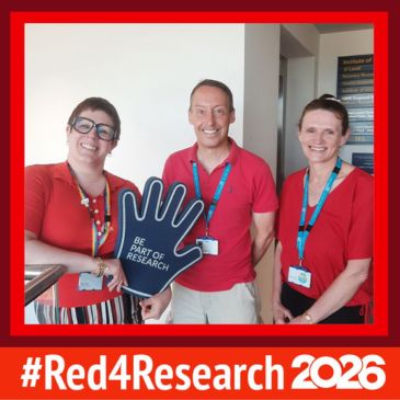£ people wearing red are smiling at the camera, one is holding a large blue foam hand, text reads red 4 research 2026