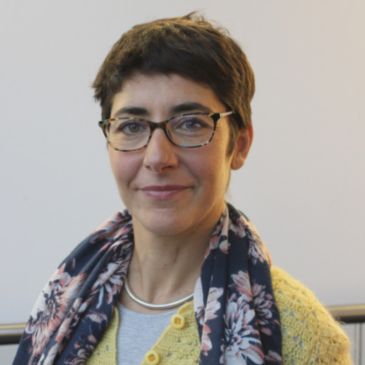 A person (Elvira) wearing glasses and a scarf is smiling at the camera