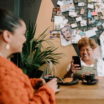 Two people are sitting at a table, one is holding a mobile phone. There's a plant in the background and a notice board 