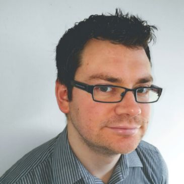 A person wearing glasses (Paul Briley) is smiling at the camera