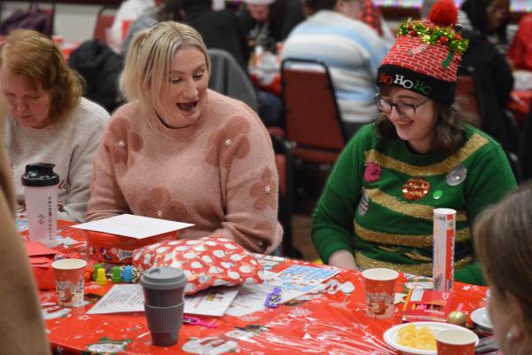 Group of people seated around a table covered with red holiday-themed wrapping paper and craft supplies, engaging in festive bingo.