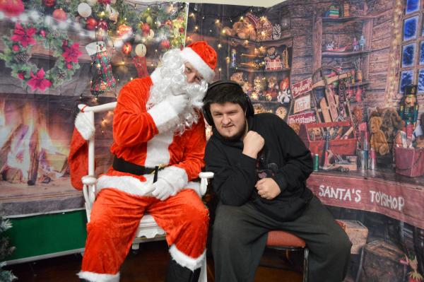 Father Christmas sitting next to a Sam in a cosy, decorated grotto with Christmas lights.