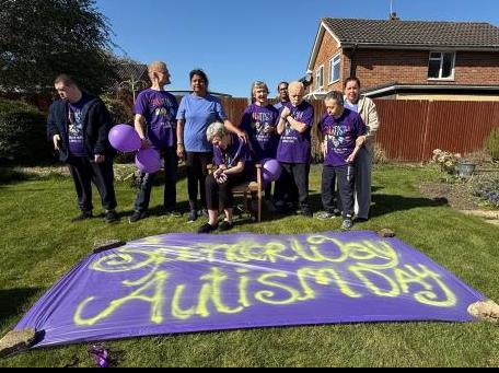 A group of people we support stand in front of an Autism Day banner - all wearing purple.