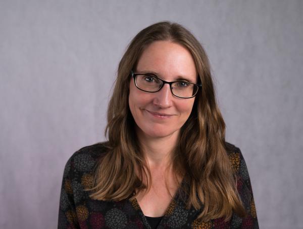 A head and shoulders photo of a smiling woman with long brown hair, dark rimmed glasses and a dark top.