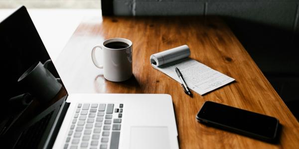 Writing materials on a desk, laptop, coffee mug, pen and paper and a smart phone